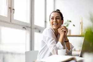 A smiling woman sitting at a desk with a laptop, cup, and books, wearing a white shirt and a bracelet, in a room with glass windows and a plant in the corner.