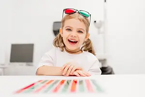 A young girl is smiling and wearing a white shirt and a pair of red and green glasses. She is sitting on a chair with her hands on the table in front of her, looking at something in front of her. The table has colorful objects on it. Behind her is a blurry background of a monitor, a desk, and a lamp.