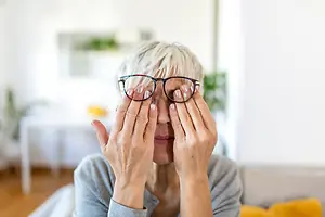 A woman sitting on a couch is wearing glasses and covering her eyes with her hands
