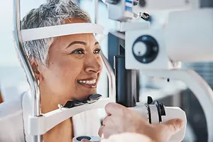 An older woman with short gray hair is having her eyes examined by a doctor or optometrist using an ophthalmoscope