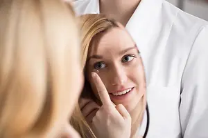 A woman with blonde hair is looking at herself in the mirror while wearing a contact lens.
