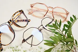 Three pairs of glasses are displayed on a white surface with plants, one pair has a pink frame and the other two pairs have brown frames.