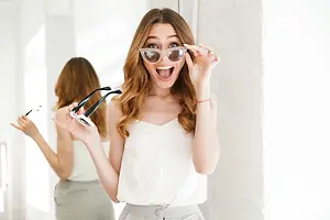 A woman with long brown hair is smiling and holding a pair of glasses in front of a mirror