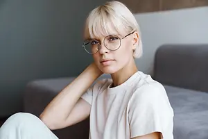A woman with short blonde hair wearing glasses and a white t-shirt sitting on a couch