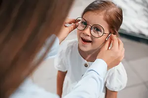 A young girl wearing glasses is smiling and looking at something while a woman adjusts her glasses.