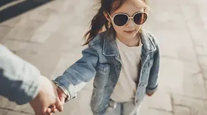A young girl wearing sunglasses and a denim jacket is holding hands with a person on a sidewalk.