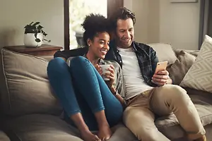 A man and woman are sitting on a couch smiling and holding a phone and a cup