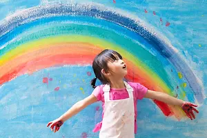 A young girl is standing with her arms outstretched in front of a rainbow painted on a wall.