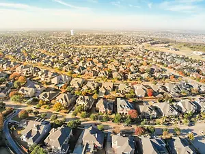 An aerial view of a residential neighborhood with houses, trees, and roads under a blue sky with some clouds.