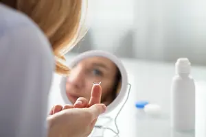 A woman is adjusting a contact lens in a mirror with a bottle of solution nearby.