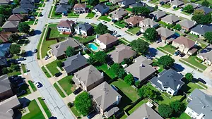 A top-down view of a neighborhood with houses and cars parked on the side streets and driveways. The houses are mostly two-story and have well-kept lawns and driveways. There are trees and a pool in the neighborhood.