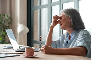 An elderly woman is sitting at a desk and rubbing her eyes while looking at a laptop. Behind her is a window with a plant and a lamp.