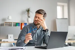 A man is sitting in front of a desk with his hand on his forehead and a laptop in front of him.