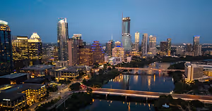 An aerial view of a city skyline at dusk with high-rise buildings, a bridge over a river, and illuminated streets.