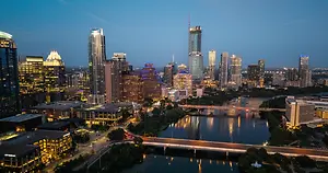 An aerial view of a city skyline at dusk with high-rise buildings, a bridge over a river, and illuminated streets.