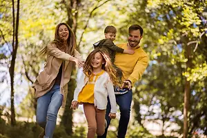 Four people, possibly a family, are running in the forest with smiling faces on a sunny day.