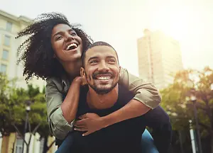 A man is carrying a woman on his back in a park while they are both smiling.