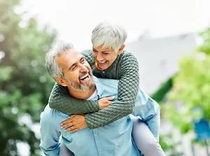 An elderly man with gray hair is carrying his white-haired wife on his shoulders while both are smiling in a park setting.