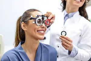 A smiling woman wearing eye glasses is getting an eye exam from a female doctor in a medical office