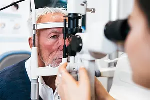 An old man wearing a ring is looking into an ophthalmoscope being held by a woman.