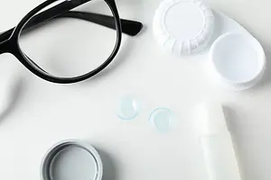 A pair of glasses, a lens case with a lens, and a bottle of lens solution are placed on a white surface.