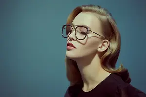 A woman wearing glasses and a black top is looking upwards with her eyes closed