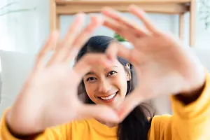 A woman is making a heart shape with her hands and smiling in a room.