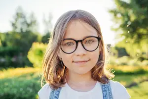 A young girl wearing glasses is smiling and standing in a park on a sunny day.