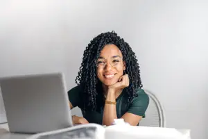 A woman in a green shirt sits at a desk with a laptop, smiling and posing for a photo.