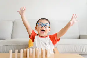 A boy in a white and orange shirt is smiling and raising his hands while playing with wooden blocks on a table in a room with a couch and a white wall