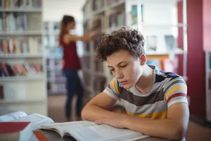 A young boy in a library is reading a book while a woman in a red dress stands behind him near a bookshelf.