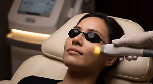 A woman is sitting on a chair, wearing black goggles, and receiving a facial treatment with a laser device.