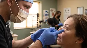 A doctor wearing a mask and blue gloves is examining a woman's eyes using a magnifying glass in a clinic