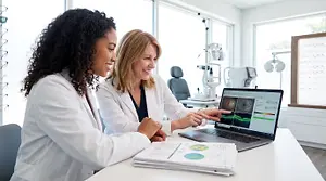 Two female optometrists in white coats reviewing patient files and diagnostic images on a laptop in a clinic