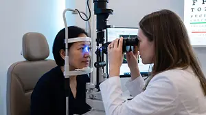 A patient at a contact lens fitting follow-up visit, optometrist reviewing lens fit under a slit lamp in a warm modern clinic