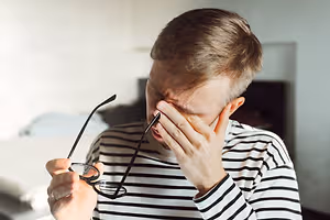 A man wearing a striped shirt holds glasses while rubbing his eye, possibly indicating discomfort or needing adjustment.