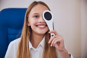 A young girl with long blonde hair is holding a white eye chart with a black circle in front of her face, smiling.