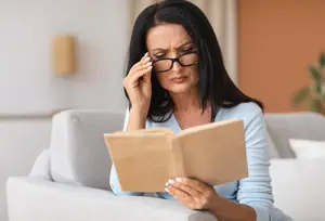 An older woman with glasses is sitting on a couch, holding a book, and has her hand on her forehead.