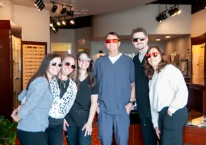 A group of people are standing together and smiling for a picture, they are wearing sunglasses, the man in the middle is wearing a watch, they are in a room with glass cabinets, a potted plant, and ceiling lights.
