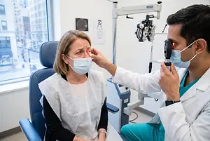 A doctor in a white coat and mask is examining a patient's eye using an ophthalmoscope in a clinic setting