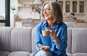 A woman with gray hair sits on a couch, smiling while holding a coffee cup and saucer.