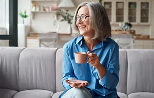 A woman with gray hair sits on a couch, smiling while holding a coffee cup and saucer.