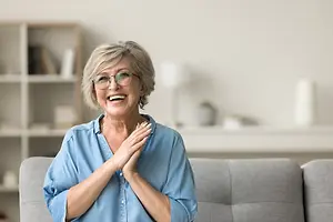 Elderly woman with glasses sitting on a couch smiling and posing for the camera.