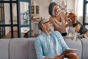 An elderly couple sitting on a couch with their dog while the woman is petting the dog and the man is smiling while holding a tablet.