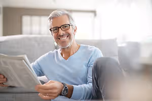 A man wearing glasses and a blue shirt is sitting on a couch and holding a newspaper, smiling while looking to his left in a room with white walls and large windows. 