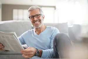 A man wearing glasses and a blue shirt is sitting on a couch and holding a newspaper, smiling while looking to his left in a room with white walls and large windows. 