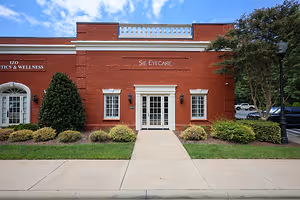 The front entrance of Sie Eyecare, a medical clinic with a red brick exterior, a white arched entrance, glass doors, and bushes in front.