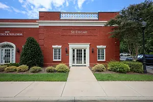 The front entrance of Sie Eyecare, a medical clinic with a red brick exterior, a white arched entrance, glass doors, and bushes in front.
