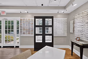 The interior of a modern optical shop features a black door with four windows, a white table, a black desk with a drawer, and a wall adorned with various eyeglass frames. The shop has wooden flooring and glass windows, and an exit sign is visible above the door.