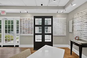 The interior of a modern optical shop features a black door with four windows, a white table, a black desk with a drawer, and a wall adorned with various eyeglass frames. The shop has wooden flooring and glass windows, and an exit sign is visible above the door.